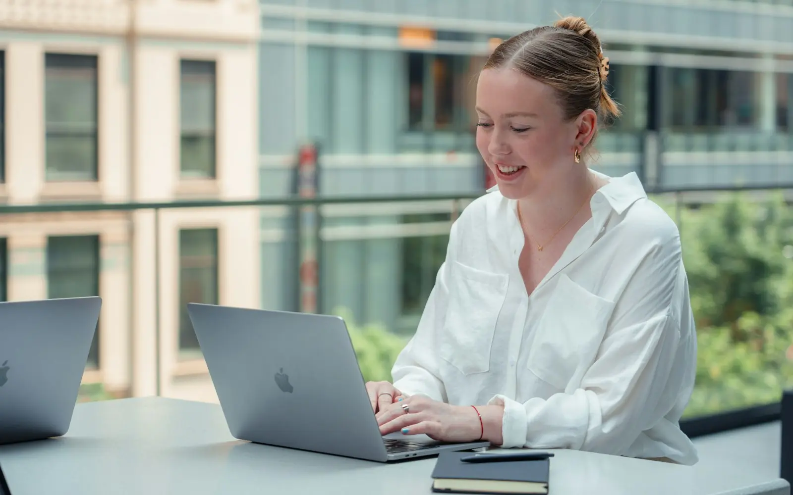 Woman smiling while working on a laptop at a table by a glass office window