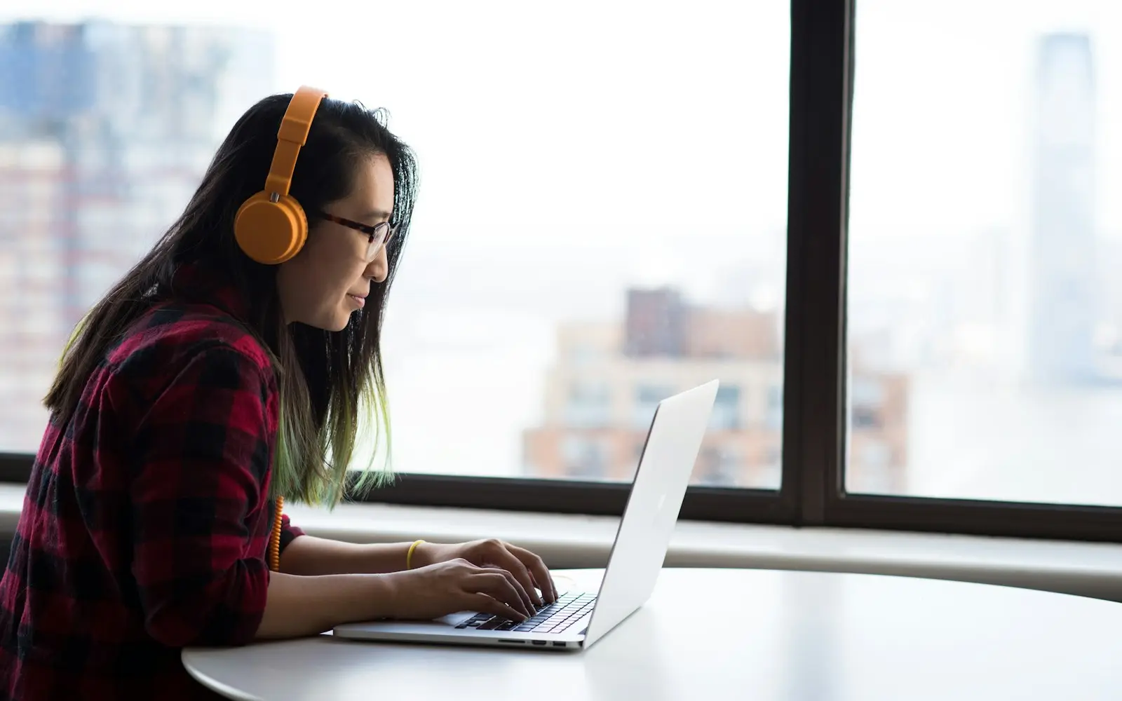 Woman with headphones working on a laptop beside a city-view window