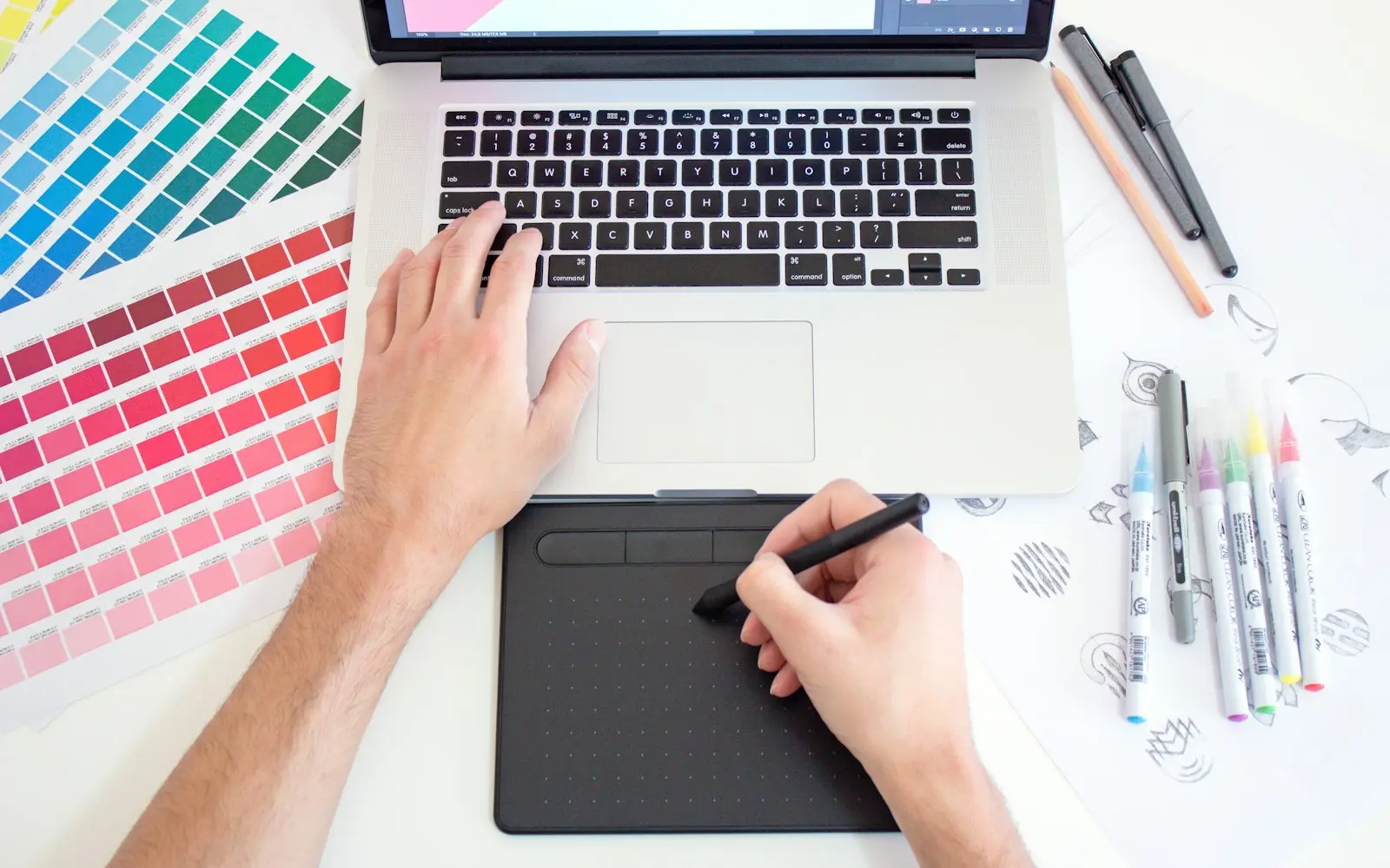 Designer's hand drawing on a graphics tablet beside a laptop and colour swatches