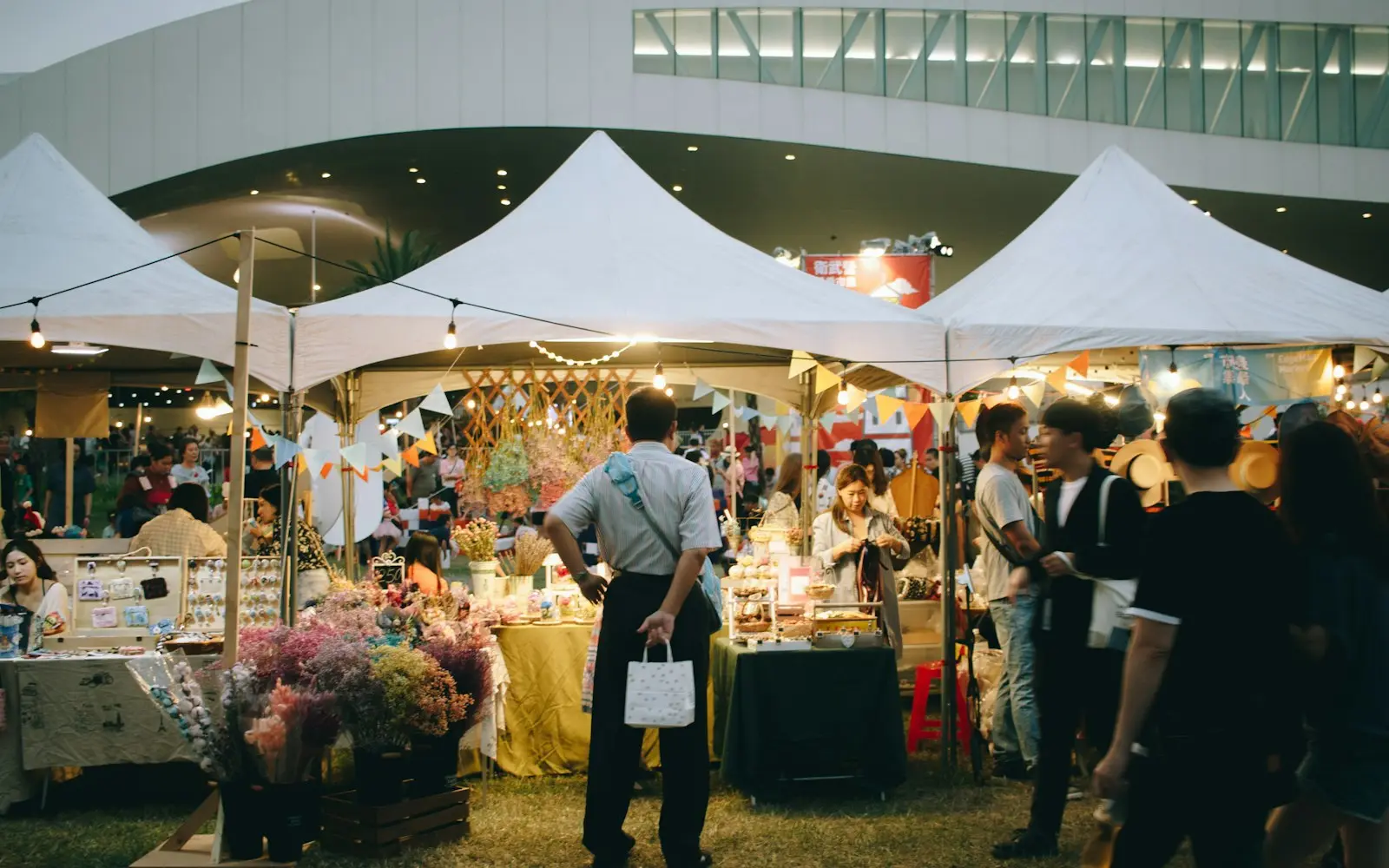 Shoppers browsing vendor stalls under white tents at an evening market