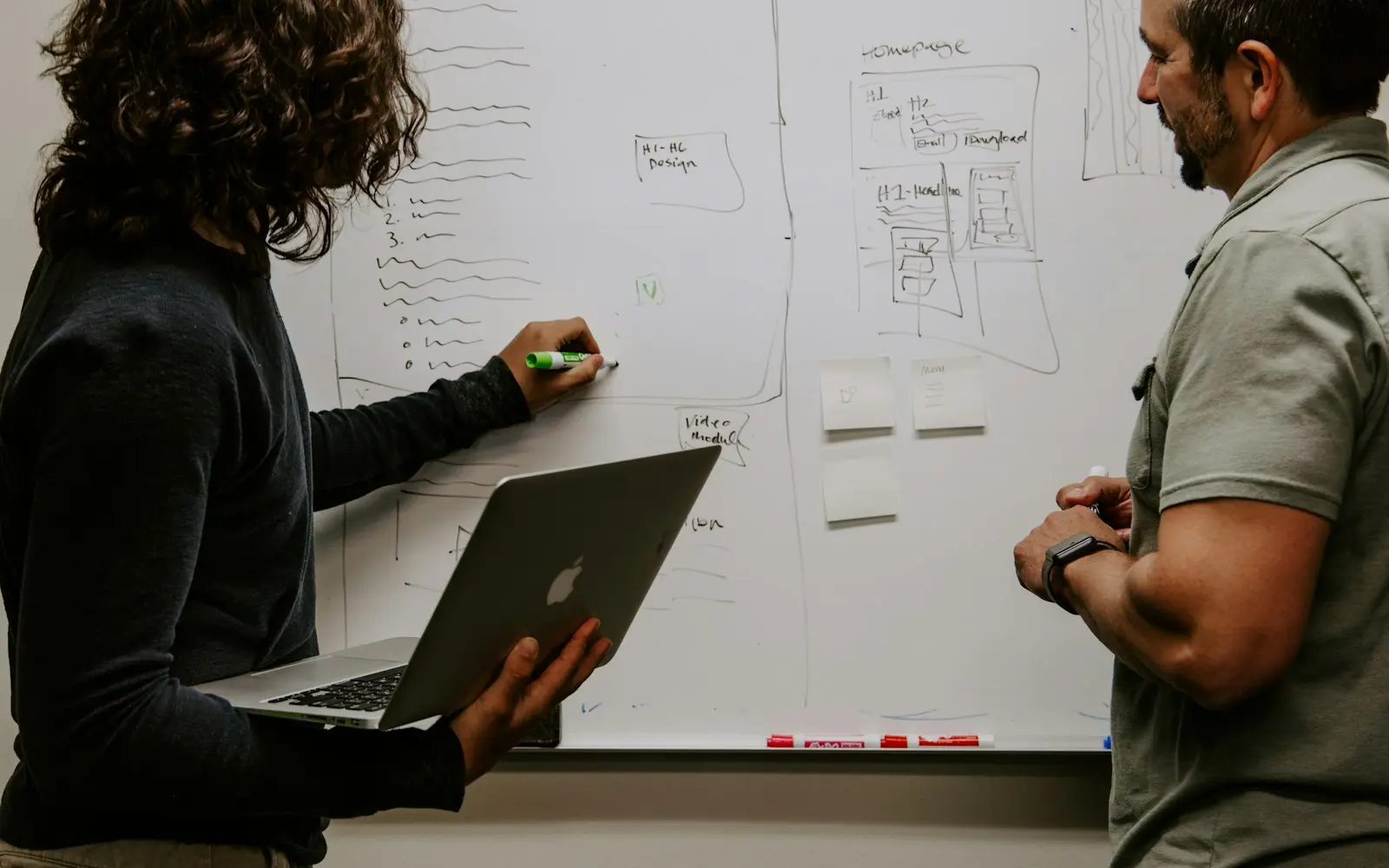 Two colleagues sketching a plan on a whiteboard, one holding a laptop