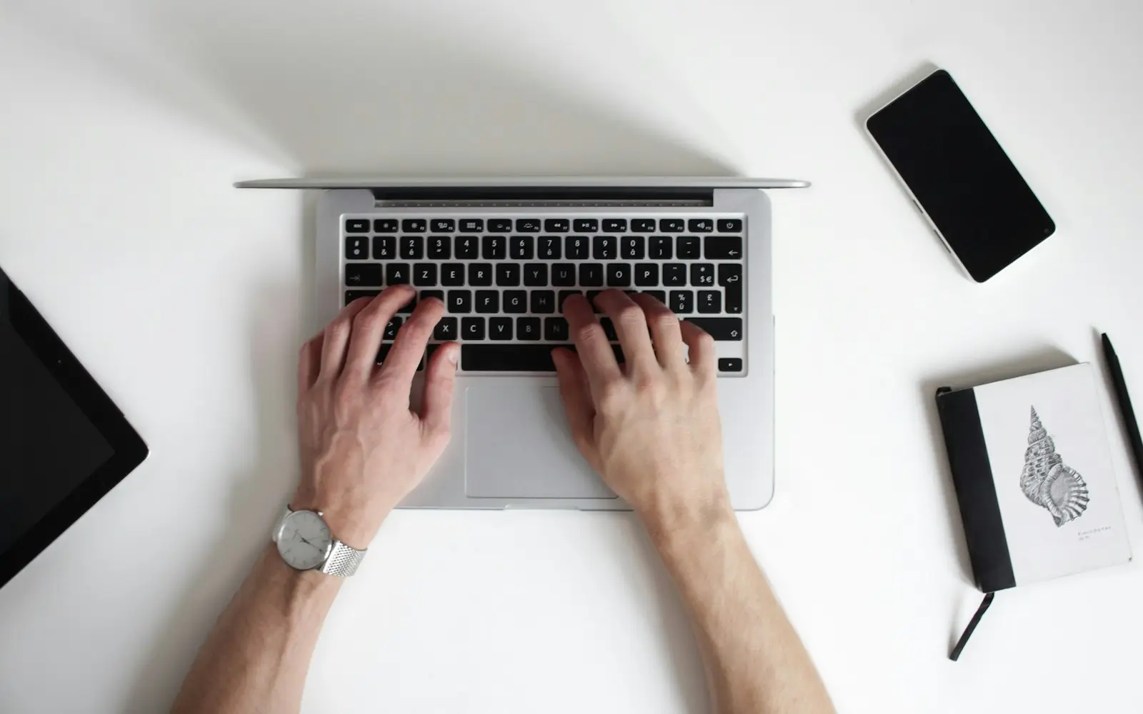 Overhead view of hands typing on a laptop beside a phone and a notebook on a white desk