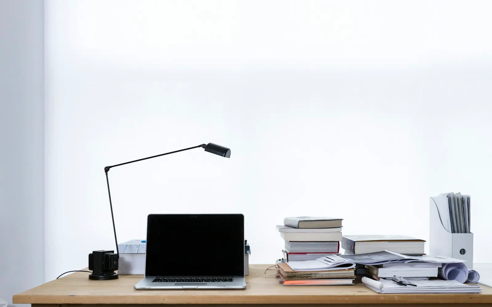 Laptop on a wooden desk beside stacked books and an articulated lamp in a white room