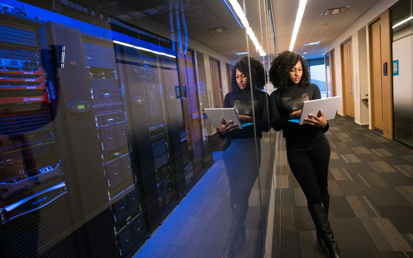Woman holding a tablet in a corridor beside glass-fronted server racks