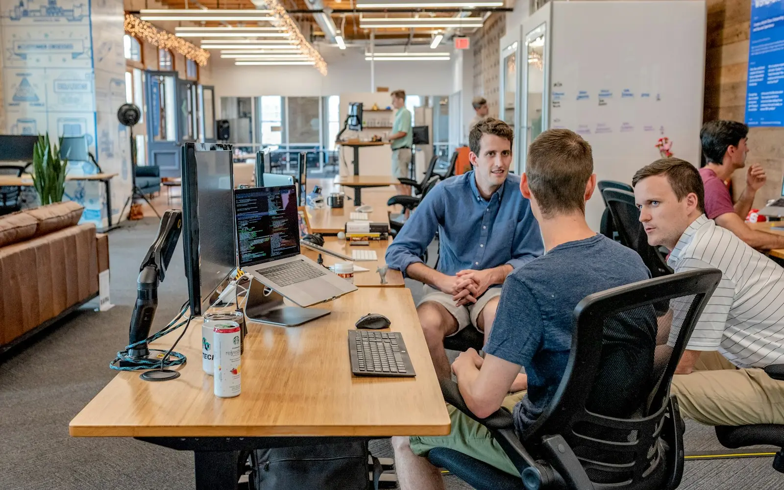 Three developers talking at a shared wooden desk with monitors in an open loft office