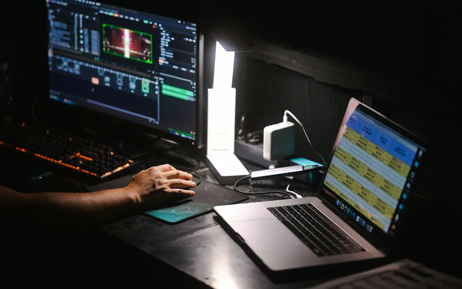 Video editing timeline on a monitor beside a laptop at a dark workstation